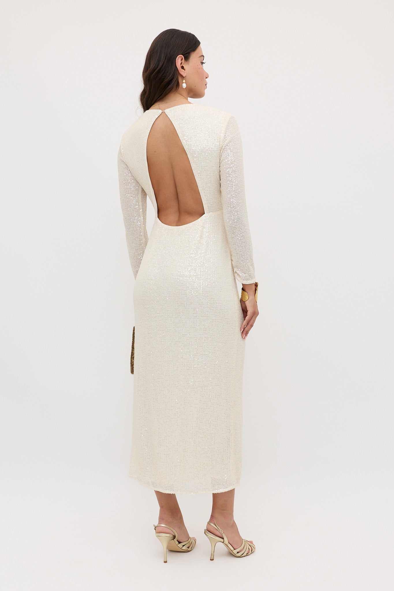 A woman with wavy brown hair wears the Lorenzo - Pearl dress, an off-white, long-sleeved sequined piece with an open back and keyhole detail. She pairs it with gold earrings and strappy heels against a plain white background.