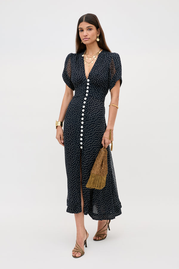 A woman in the Novara - Painted Spot Deep Navy vintage dress with white buttons, curved sleeves, gold accessories, and strappy heels holds a brown fringed handbag against a plain light background.