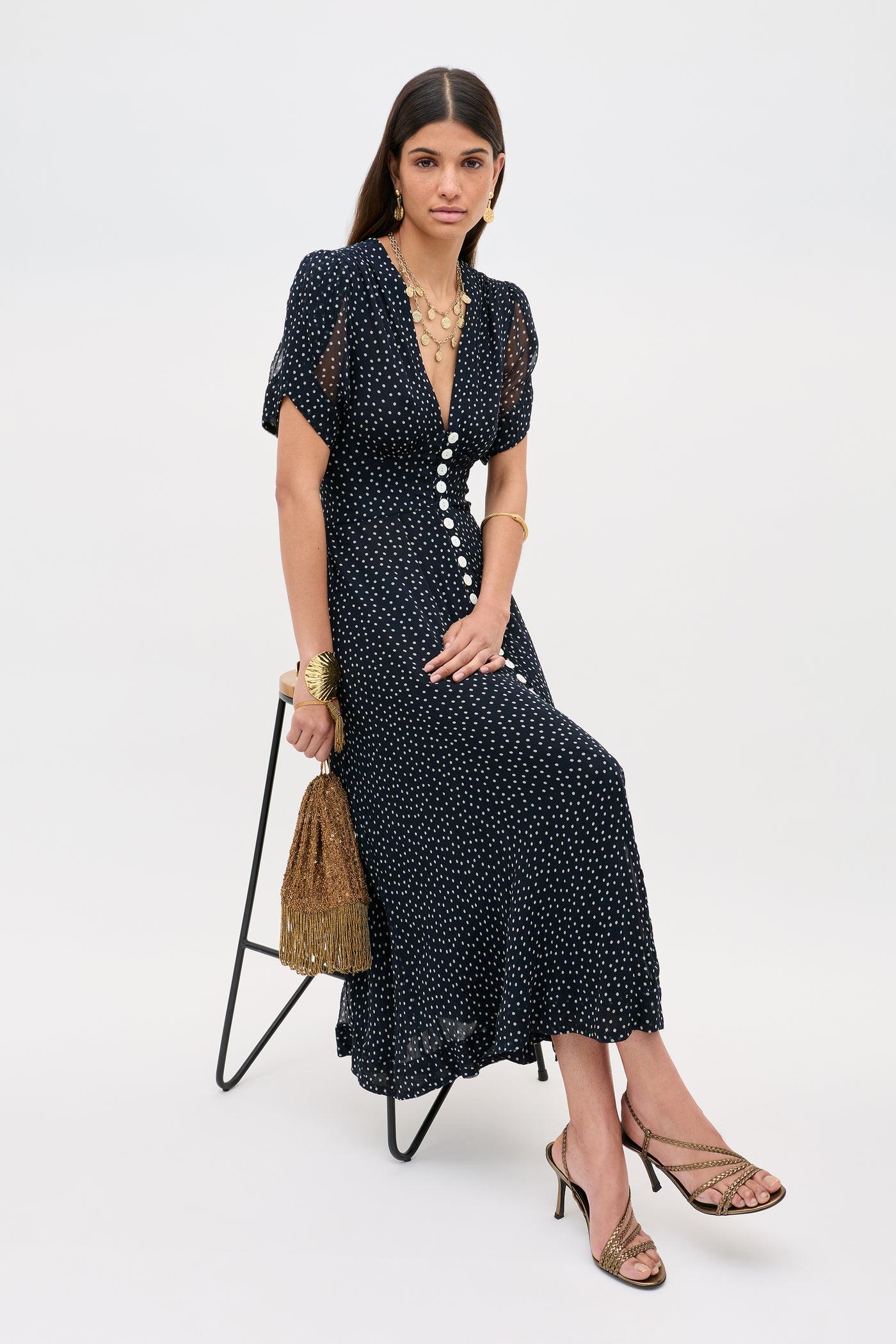 A woman sits on a stool wearing the Novara - Painted Spot Deep Navy dress, paired with gold jewelry and strappy heels. She holds a brown fringed handbag and looks at the camera against a plain light background.