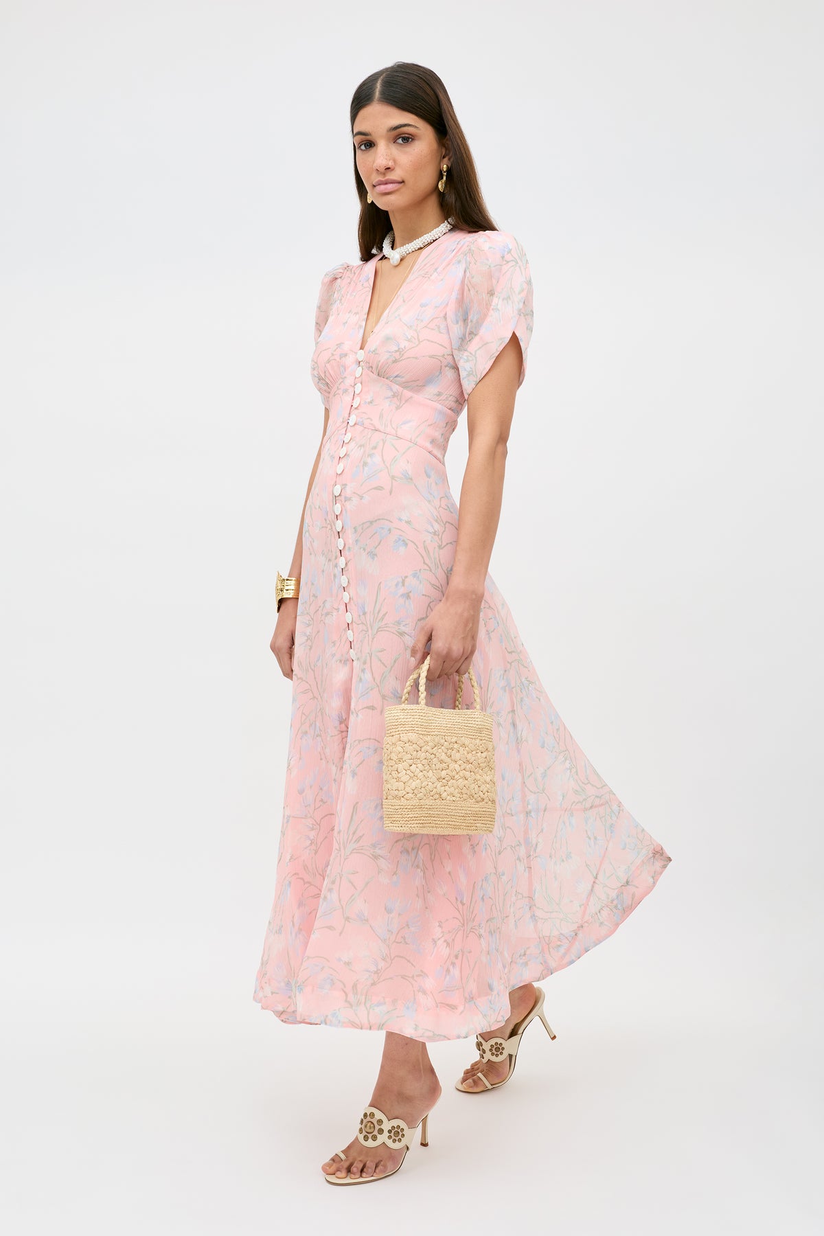 A woman stands against a plain white background wearing the Novara - Watercolour Floral Pink dress, holding a woven straw handbag, and accessorized with heeled sandals and gold jewelry.