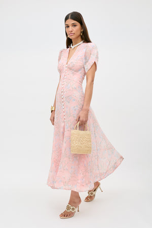A woman stands against a plain white background wearing the Novara - Watercolour Floral Pink dress, holding a woven straw handbag, and accessorized with heeled sandals and gold jewelry.