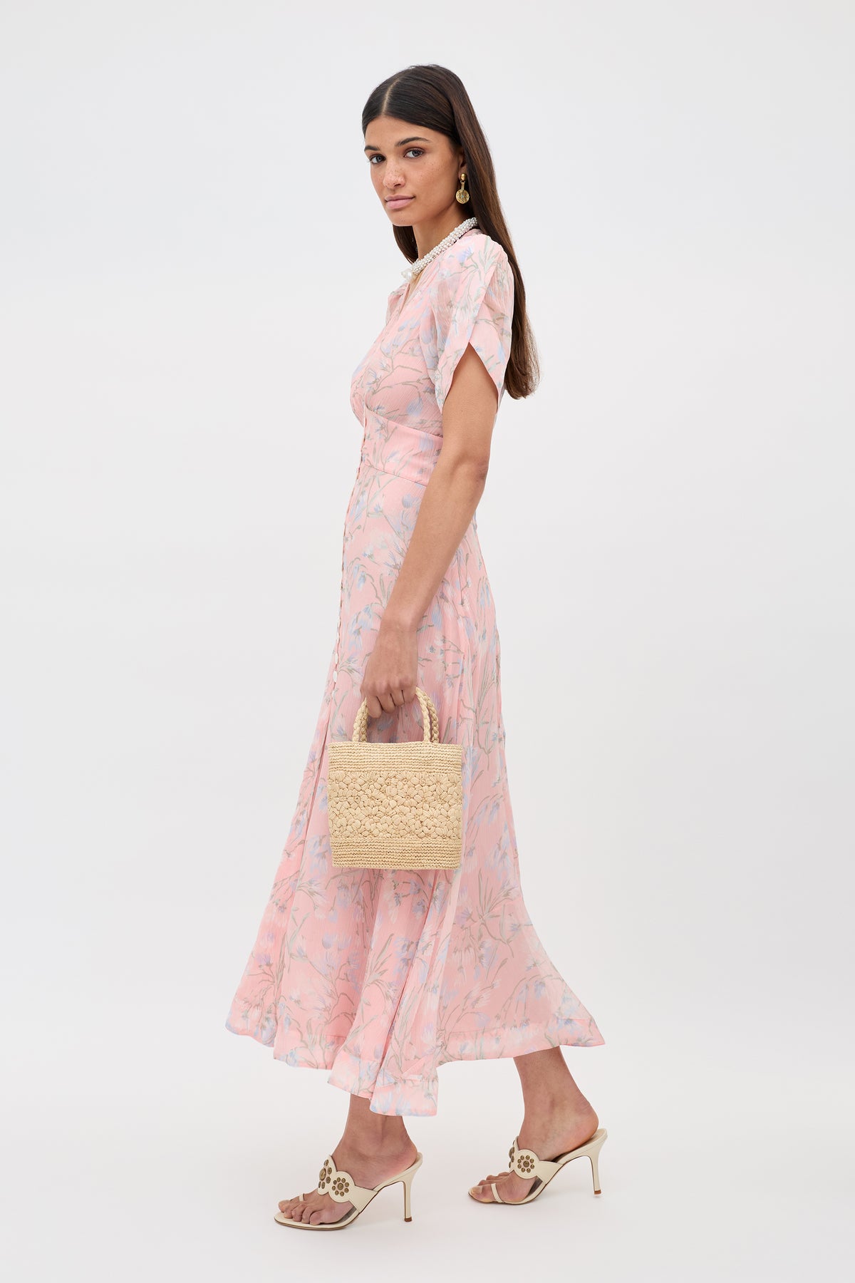 Wearing the Novara - Watercolour Floral Pink midi dress with heeled sandals and a woven straw handbag, a woman stands against a plain white background, glancing over her shoulder.