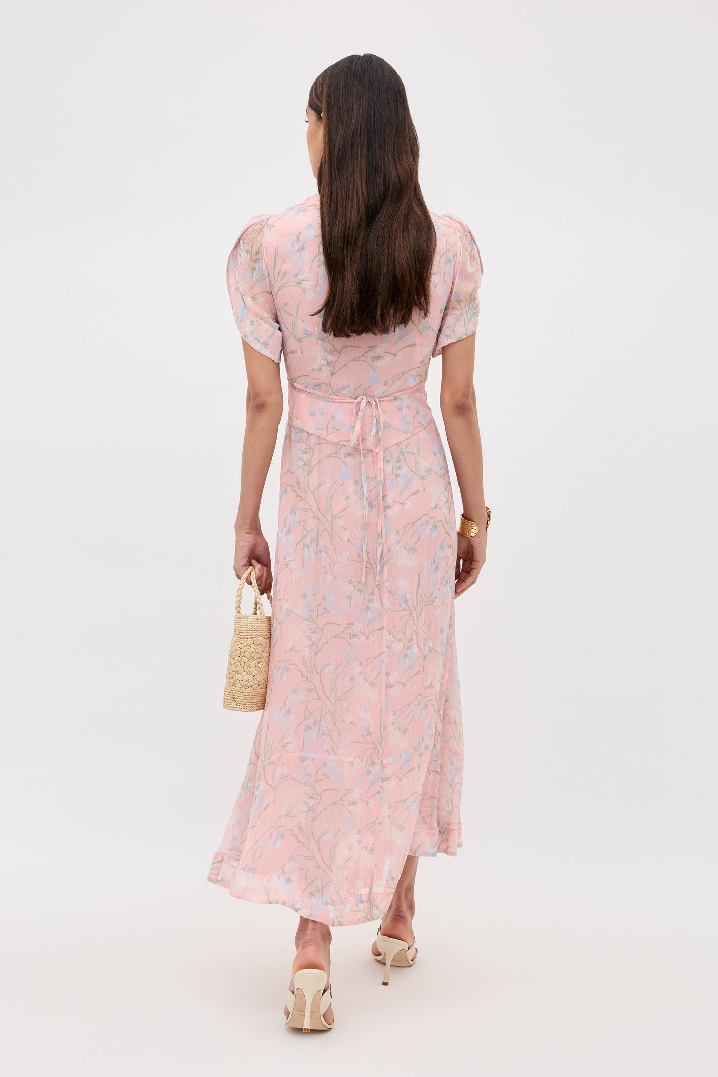 A woman with long brown hair stands facing away, wearing the Novara - Watercolour Floral Pink dress with puffed sleeves and a waist tie. She holds a small woven handbag and wears cream-heeled sandals.