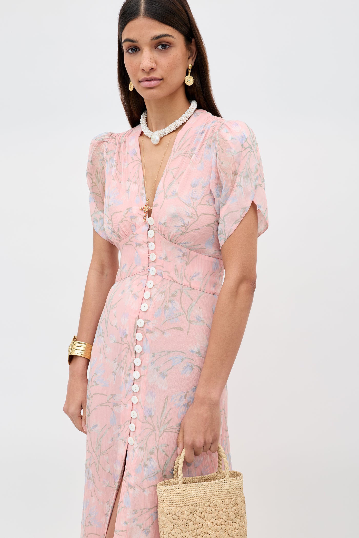 A woman in the Novara - Watercolour Floral Pink dress with white buttons holds a woven straw bag, accessorized with gold earrings, a gold bracelet, and a pearl necklace. Her straight dark hair is parted in the middle.