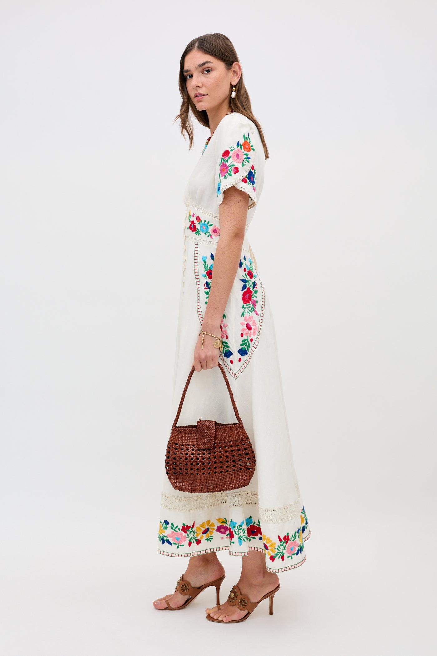 A woman wears the Novara - Anemone Embroidery Cream dress, holding a woven brown handbag. She stands in profile with tan high-heeled sandals and gold earrings against a plain white background.