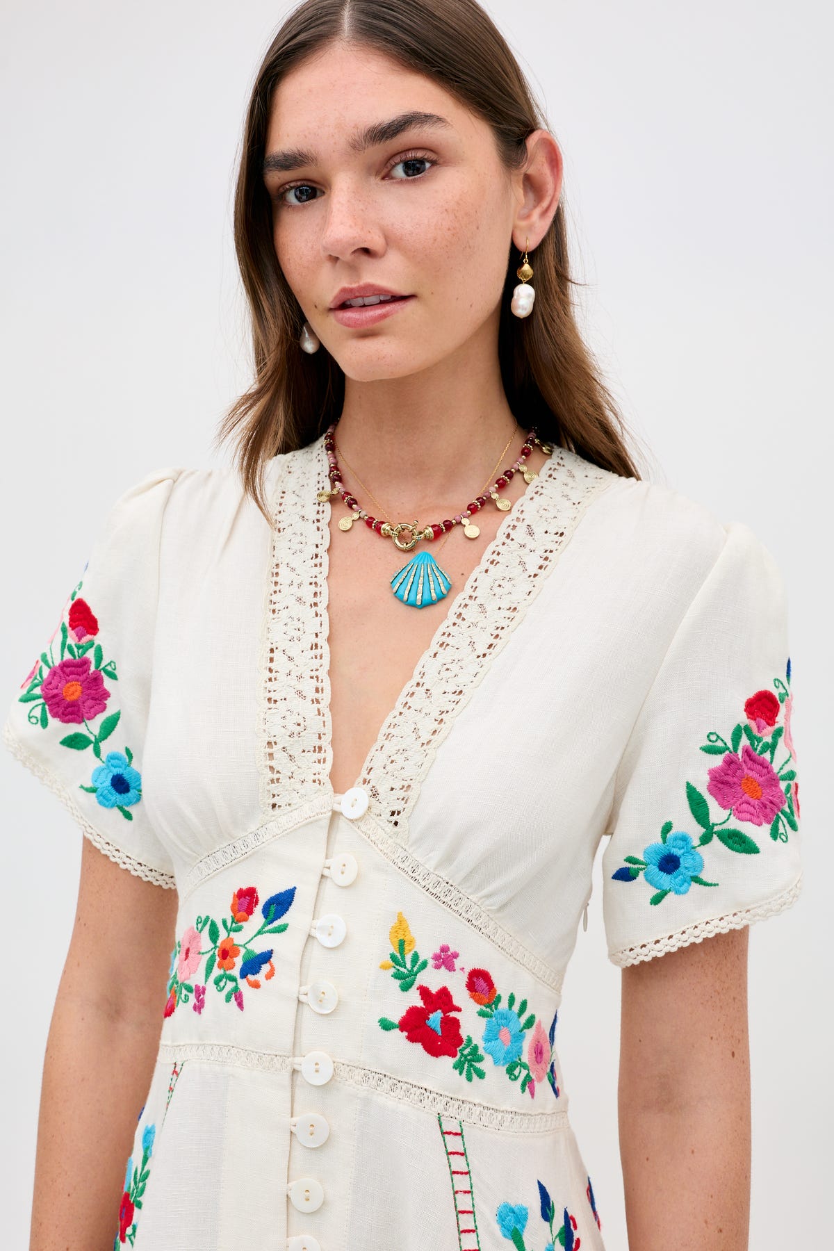 A woman wears the Tala - Burgundy dress, featuring floral embroidery and lace trim, standing against a plain background. She has long brown hair and accessorizes with pearl earrings and a blue pendant necklace.
