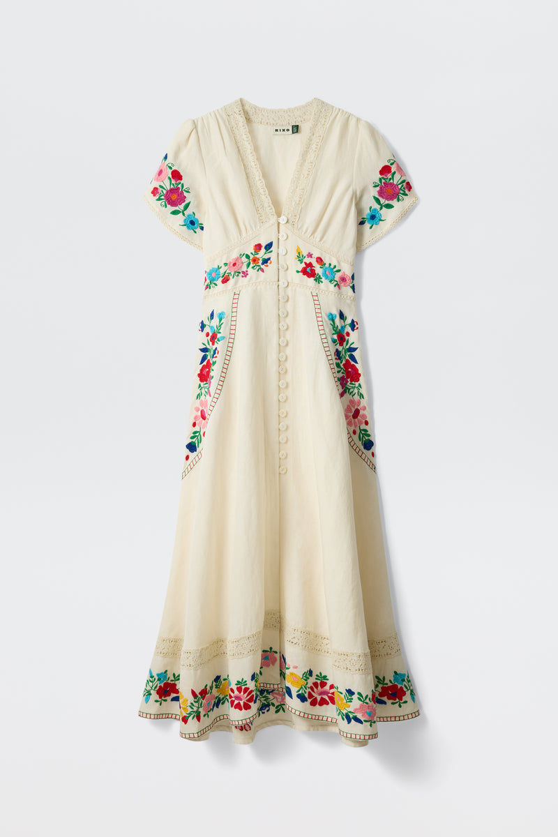 The Novara - Anemone Embroidery Cream is a cream midi dress with short sleeves, a V-neck, and decorative front buttons, featuring colorful floral embroidery on the neckline, sleeves, waist, pockets, and hem.
