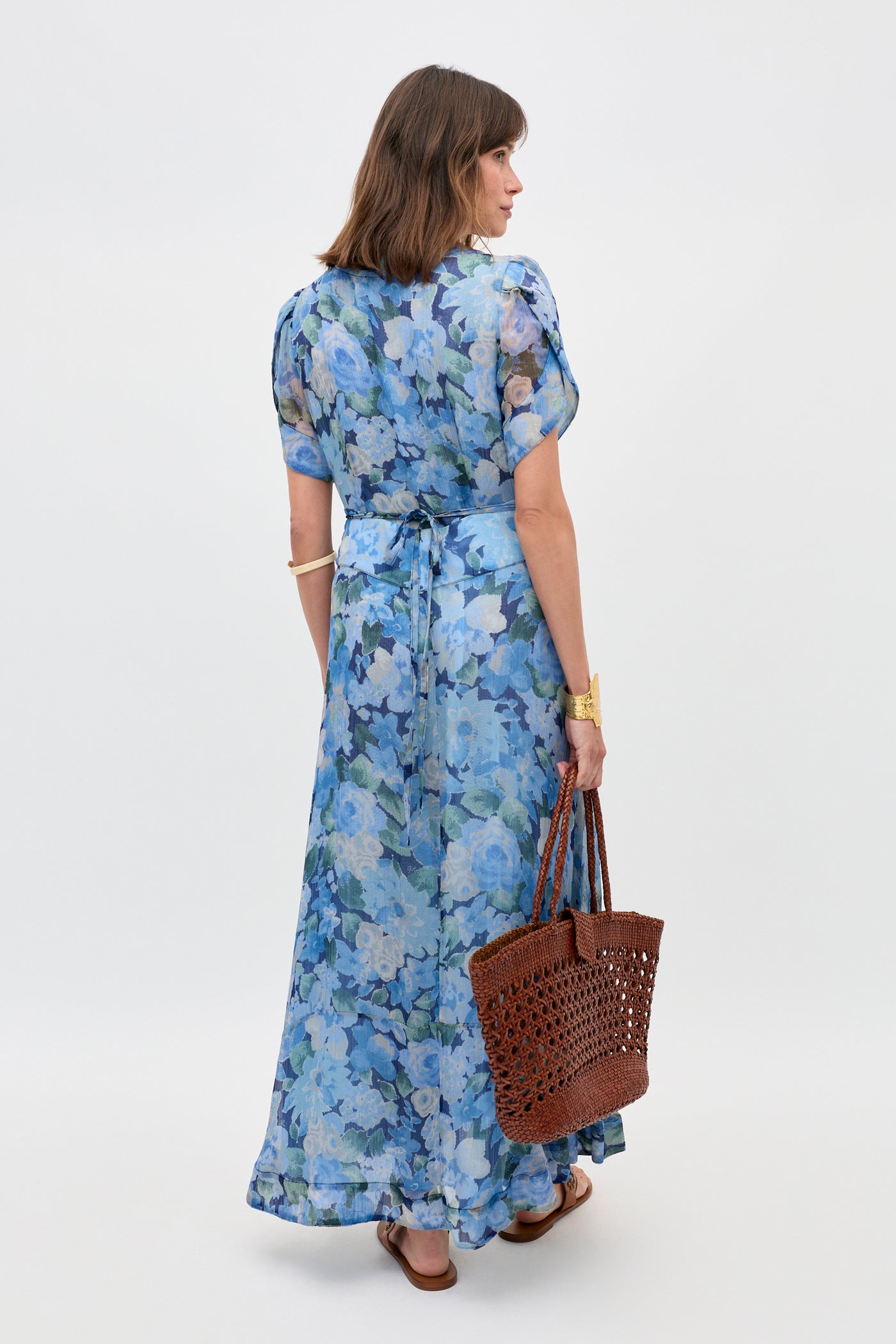 A woman faces away, wearing the Novara - Chelsea Floral Blue dress with short sleeves and a waist tie. She carries a brown woven tote, wears sandals, a gold bracelet, and a watch against a plain light background.