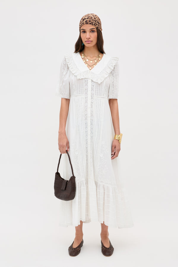 A woman stands against a plain white background wearing Edina - Pansy Embroidery White, a vintage-style midi dress with lace details and a ruffled collar, paired with a leopard-print headscarf, gold necklaces, black handbag, and brown flats.