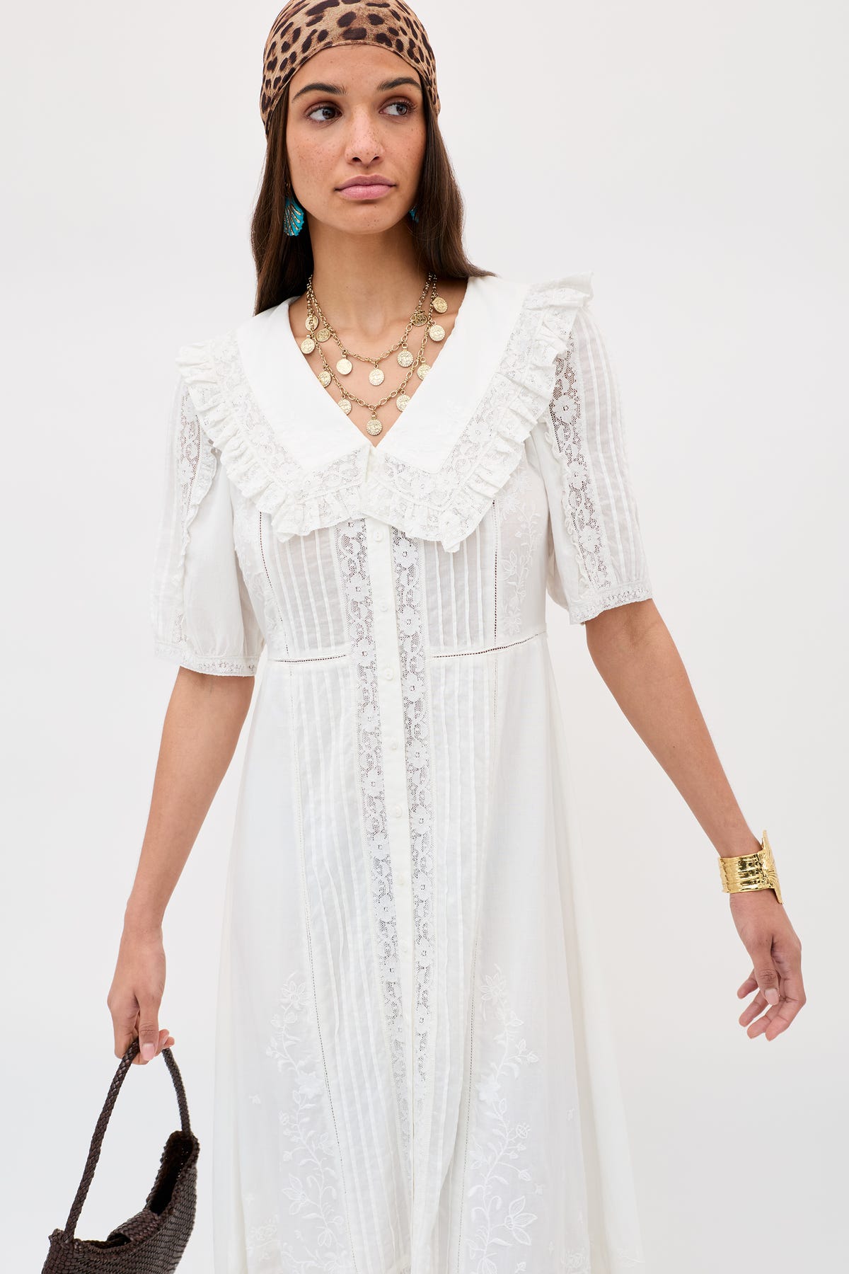 A woman wears the Edina - Pansy Embroidery White blouse with a ruffled collar, brown leopard-print headscarf, layered gold necklaces, turquoise earrings, an adjustable waist, and a brown woven handbag.
