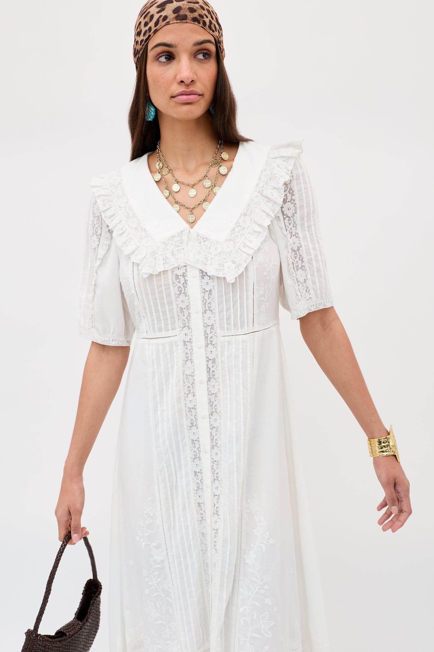 A woman wears the Edina - Pansy Embroidery White blouse with a ruffled collar, brown leopard-print headscarf, layered gold necklaces, turquoise earrings, an adjustable waist, and a brown woven handbag.