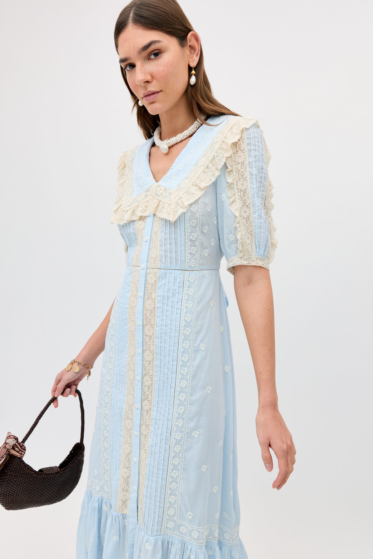 Wearing the Edina - Mira Embroidery Powder Blue dress, a woman with straight brown hair, pearl earrings, and a necklace holds a dark brown woven handbag against a plain background.