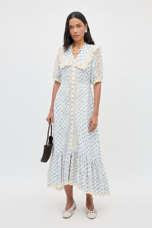 A woman stands against a white background wearing the Edina - Seville Floral Cream dress, which features delicate lace trim. She holds a small brown bag, pairs the outfit with white shoes, and completes her look with long dark hair, earrings, and a necklace.