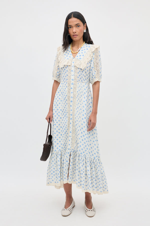 A woman stands against a white background wearing the Edina - Seville Floral Cream dress, which features delicate lace trim. She holds a small brown bag, pairs the outfit with white shoes, and completes her look with long dark hair, earrings, and a necklace.