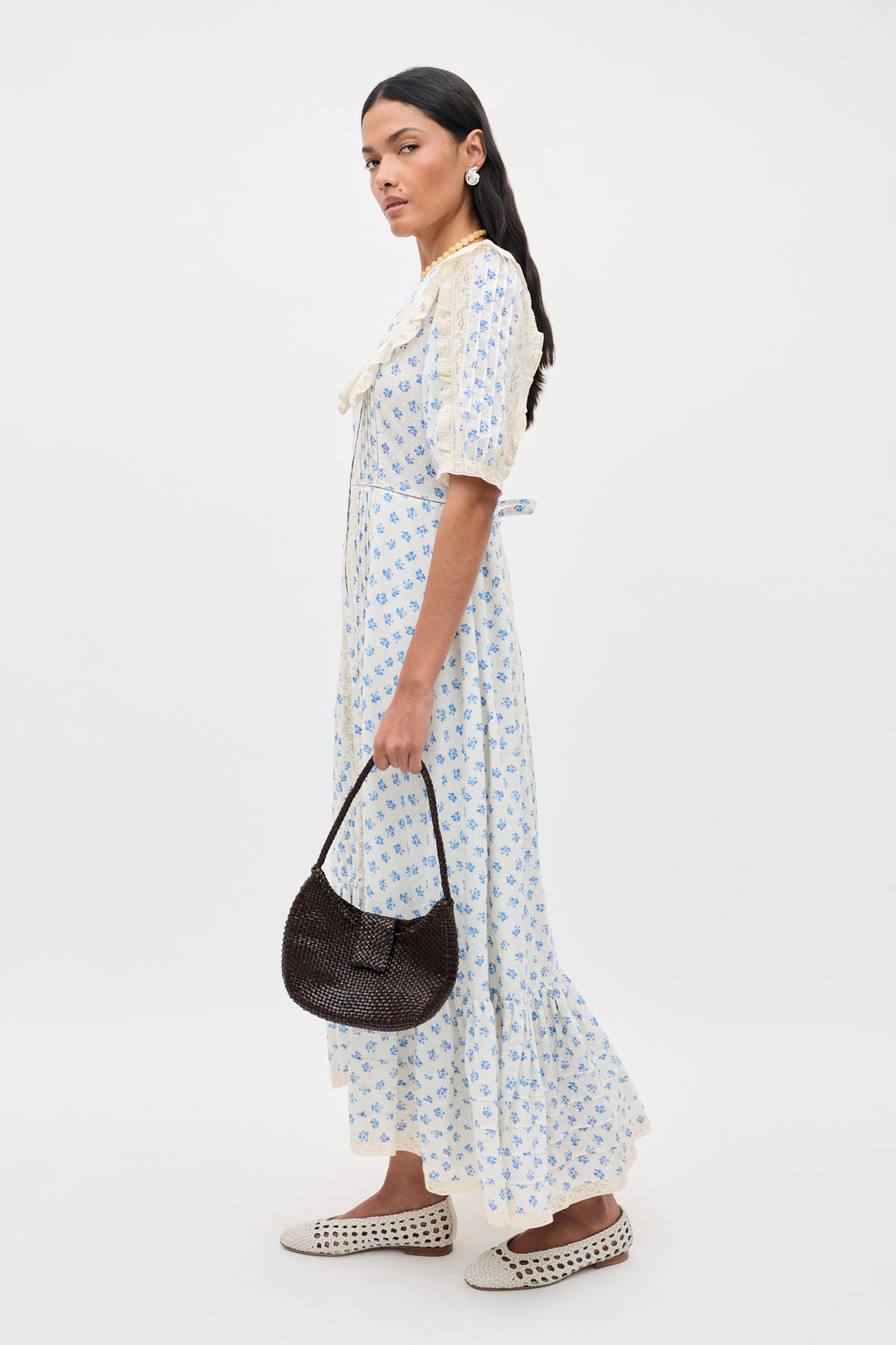 Woman wearing the Edina - Seville Floral Cream maxi dress, paired with cream woven flats and a dark brown textured handbag. She has long dark hair, pearl earrings, and is set against a neutral background.