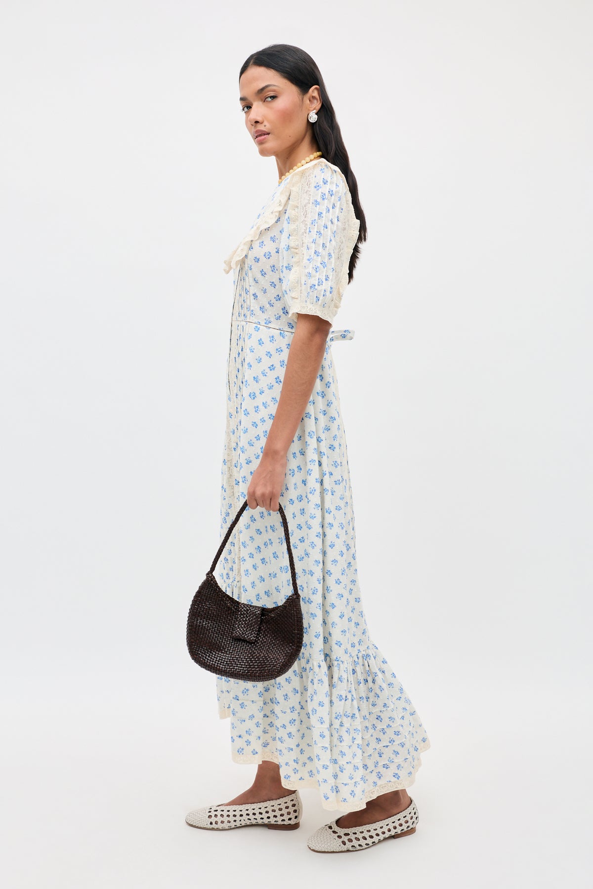 Woman wearing the Edina - Seville Floral Cream maxi dress, paired with cream woven flats and a dark brown textured handbag. She has long dark hair, pearl earrings, and is set against a neutral background.