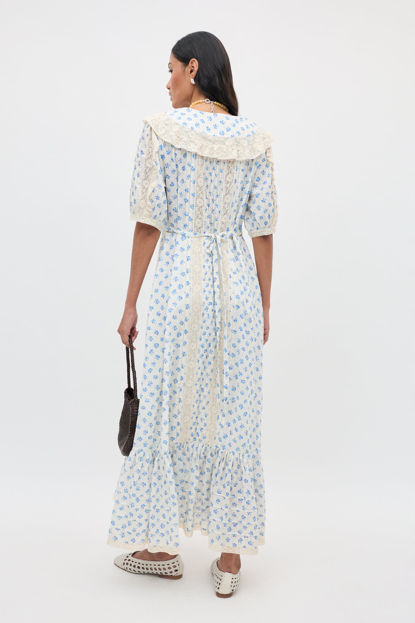 A woman stands facing away, wearing the Edina - Seville Floral Cream dress—a long, light-colored piece with blue floral patterns, lace collar, and puff sleeves. She holds a small dark bag and wears white shoes with cut-out details against a plain white background.