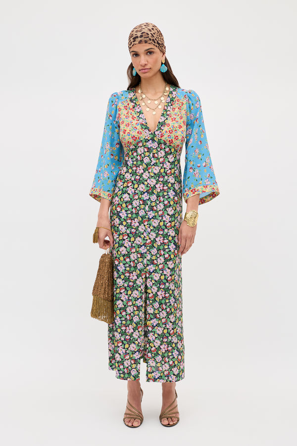 A woman in the Dahlia - Jeannie Mix Multi long floral dress with blue sleeves, layered necklaces, a gold bracelet, blue earrings, brown patterned headscarf, textured handbag, and strappy heels stands against a plain background.