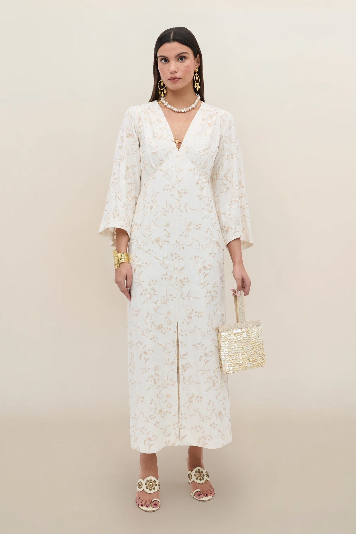 A woman stands against a plain background wearing the “Dahlia - Botanical Embroidery Pearl” dress, featuring vintage-inspired floral embroidery and flared sleeves, styled with gold jewelry, white heeled sandals, and a woven square handbag.