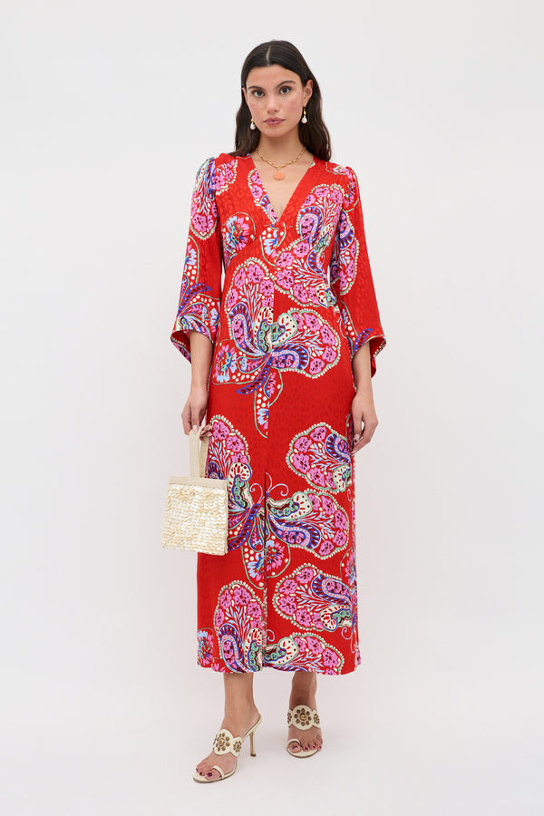 A woman with dark, wavy hair stands against a white background wearing the Dahlia - Butterfly Bloom Jacquard Red maxi dress, beige heeled sandals, and holding a small woven handbag, her expression neutral.