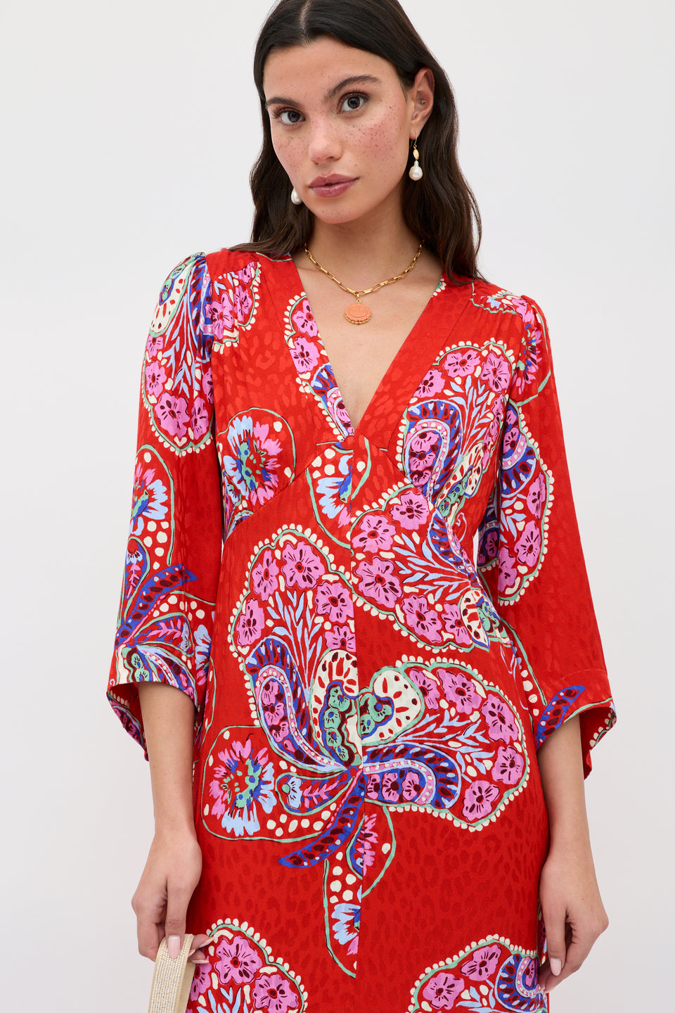 A woman wears the Dahlia - Butterfly Bloom Jacquard Red dress, featuring bold pink, purple, and blue floral patterns, a V-neckline, and three-quarter sleeves. She accessorizes with gold jewelry and a small beige bag.