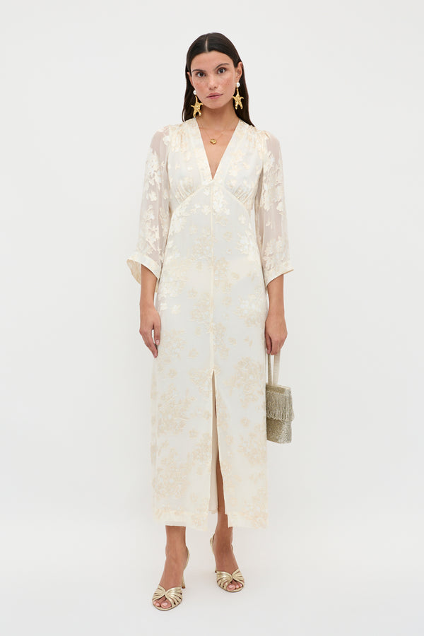 A woman poses against a white background wearing the Dahlia – Bouquet Devore Cream, an elegant ankle-length cream dress with gold floral patterns, V-neckline, and sheer sleeves. She accessorizes with a fringe handbag, star earrings, and heeled sandals.