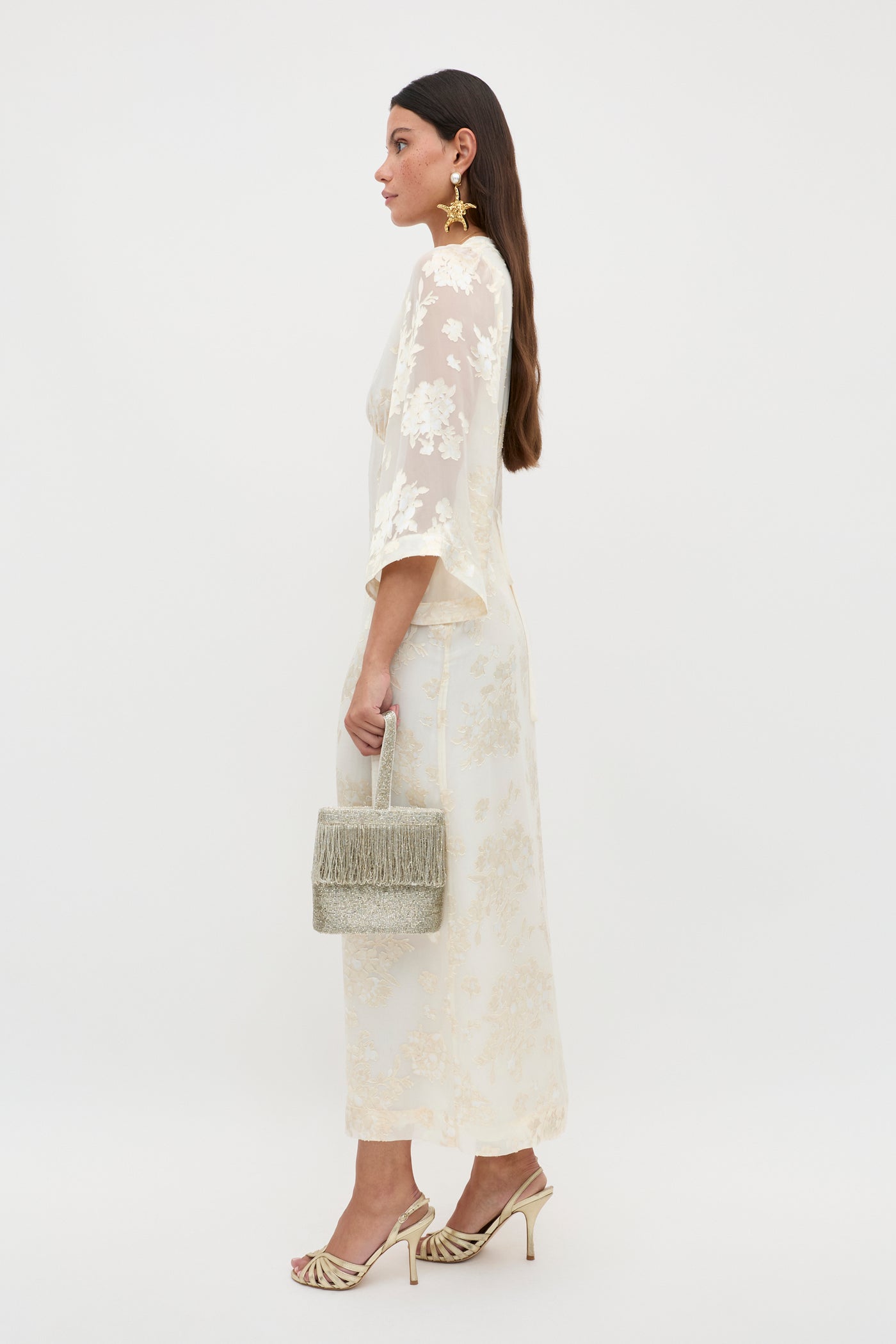 Woman wearing the Dahlia - Bouquet Devore Cream dress, holding a textured beige handbag, with gold high-heeled sandals and large gold earrings, shown in profile against a plain white background.