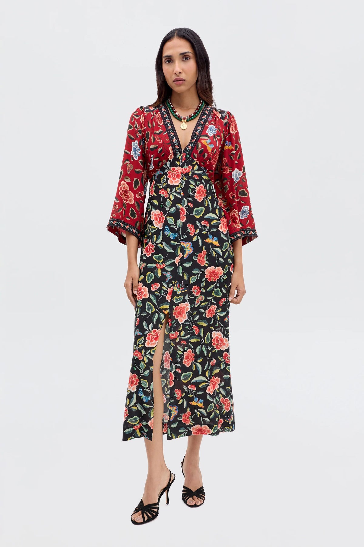 A woman wears the Dahlia - Butterfly Mix Black long-sleeved wrap dress with floral prints and red-black color block, styled with black strappy heels, making it the perfect transitional piece against a plain background.