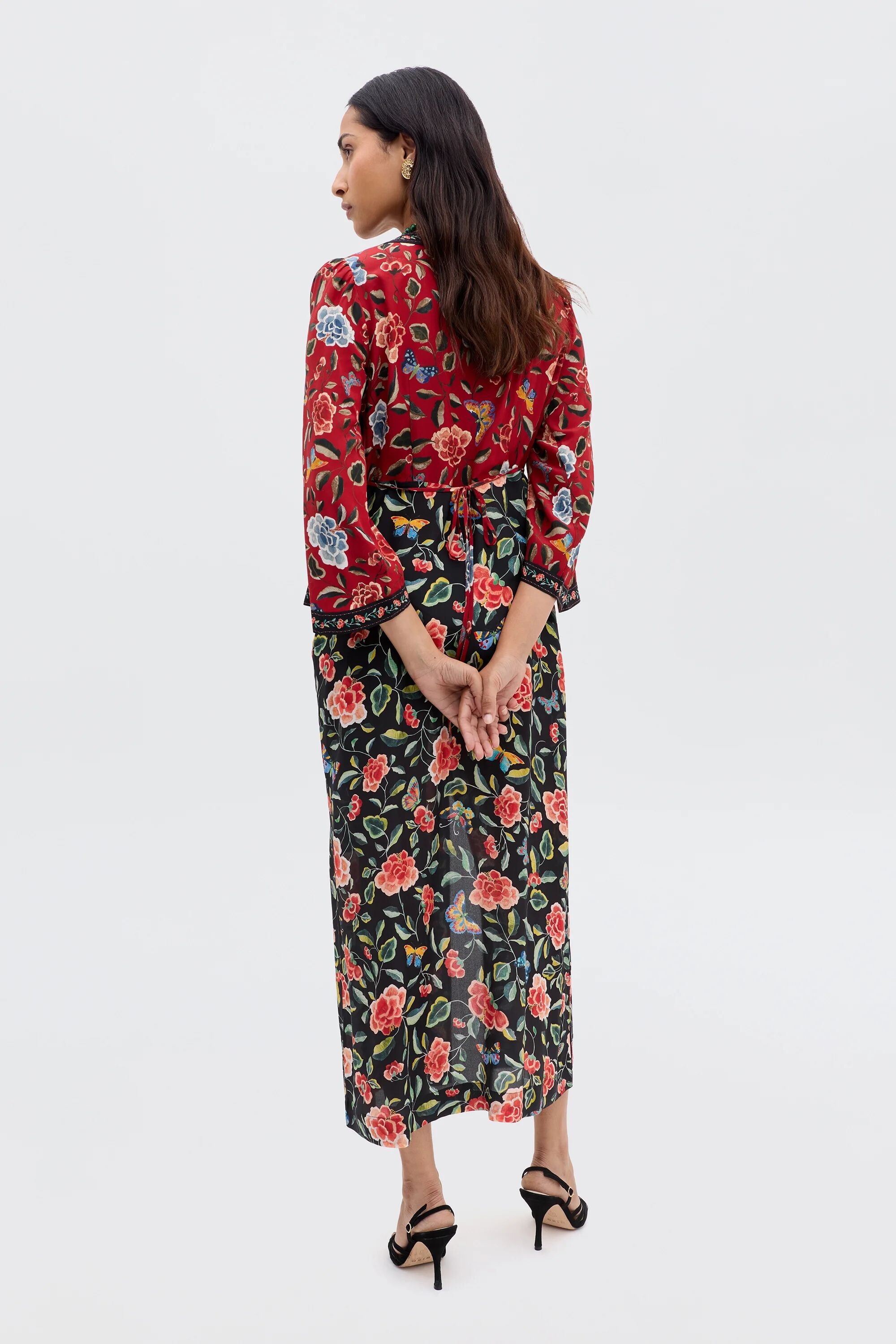A woman faces away in the Dahlia - Butterfly Mix Black, a long-sleeved, ankle-length dress with chinoiserie florals. Its red top and black skirt, styled with black heeled sandals, make it a striking transitional piece.