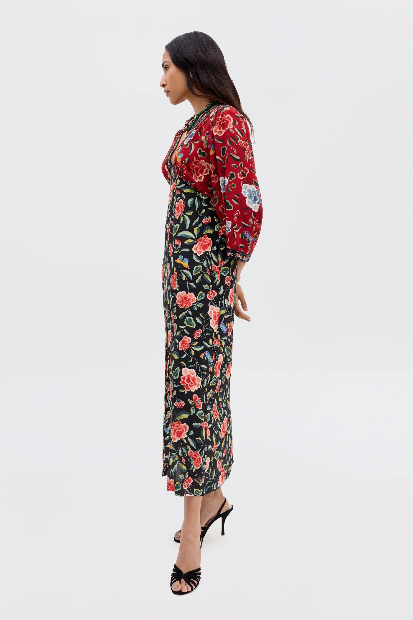 A woman stands in profile wearing the Dahlia - Butterfly Mix Black dress with chinoiserie florals, red floral-embroidered top, and black strappy high-heel sandals against a plain light background.