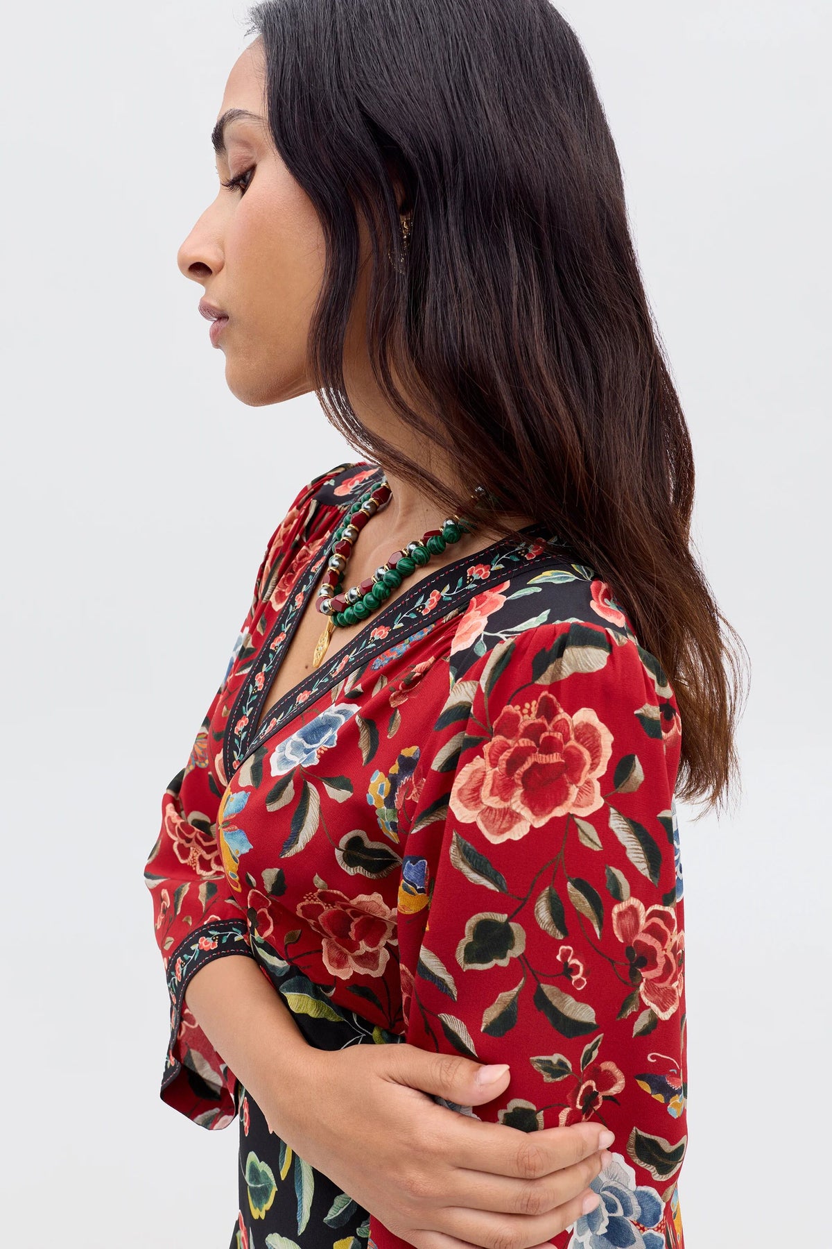 A woman with long dark hair stands in profile with arms crossed, wearing the "Dahlia - Butterfly Mix Black" dress, featuring red chinoiserie florals and a green beaded necklace, against a plain light background.