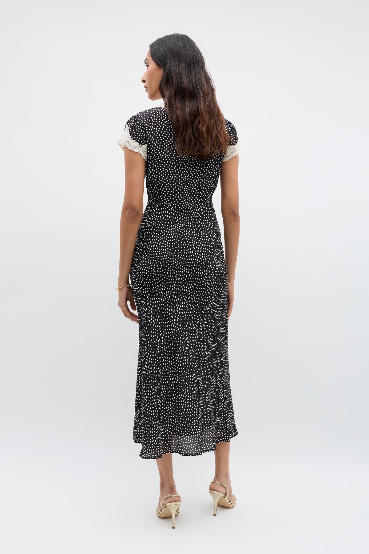 A woman with wavy brown hair faces away, wearing the Amari - Mini Polka Dot Black dress—black, short-sleeved, ankle-length with white polka dots, lace-trimmed sleeves, adjustable V-neck—and beige high heels against a light background.