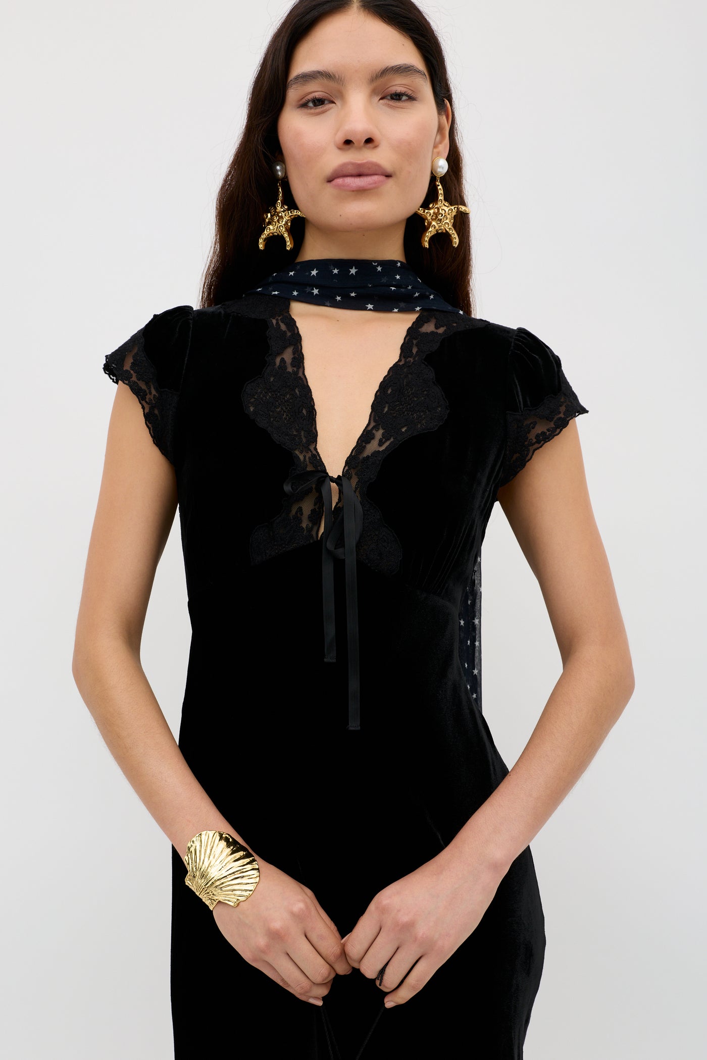 A woman wears the Amari - Black velvet evening dress with lace details, paired with a matching black scarf with white stars, gold seashell earrings, and a large shell-shaped gold bracelet.