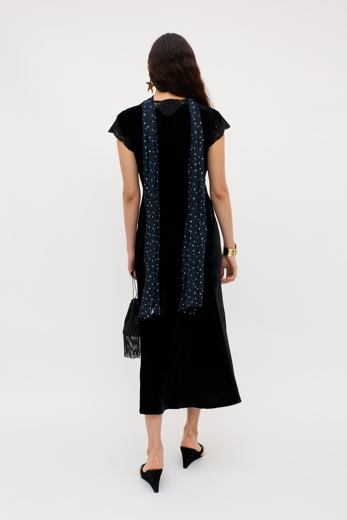A woman with loose hair wears the Amari - Black velvet dress with lace sleeves, a long blue patterned scarf, black pointed heels, a black fringed handbag, and a gold watch.