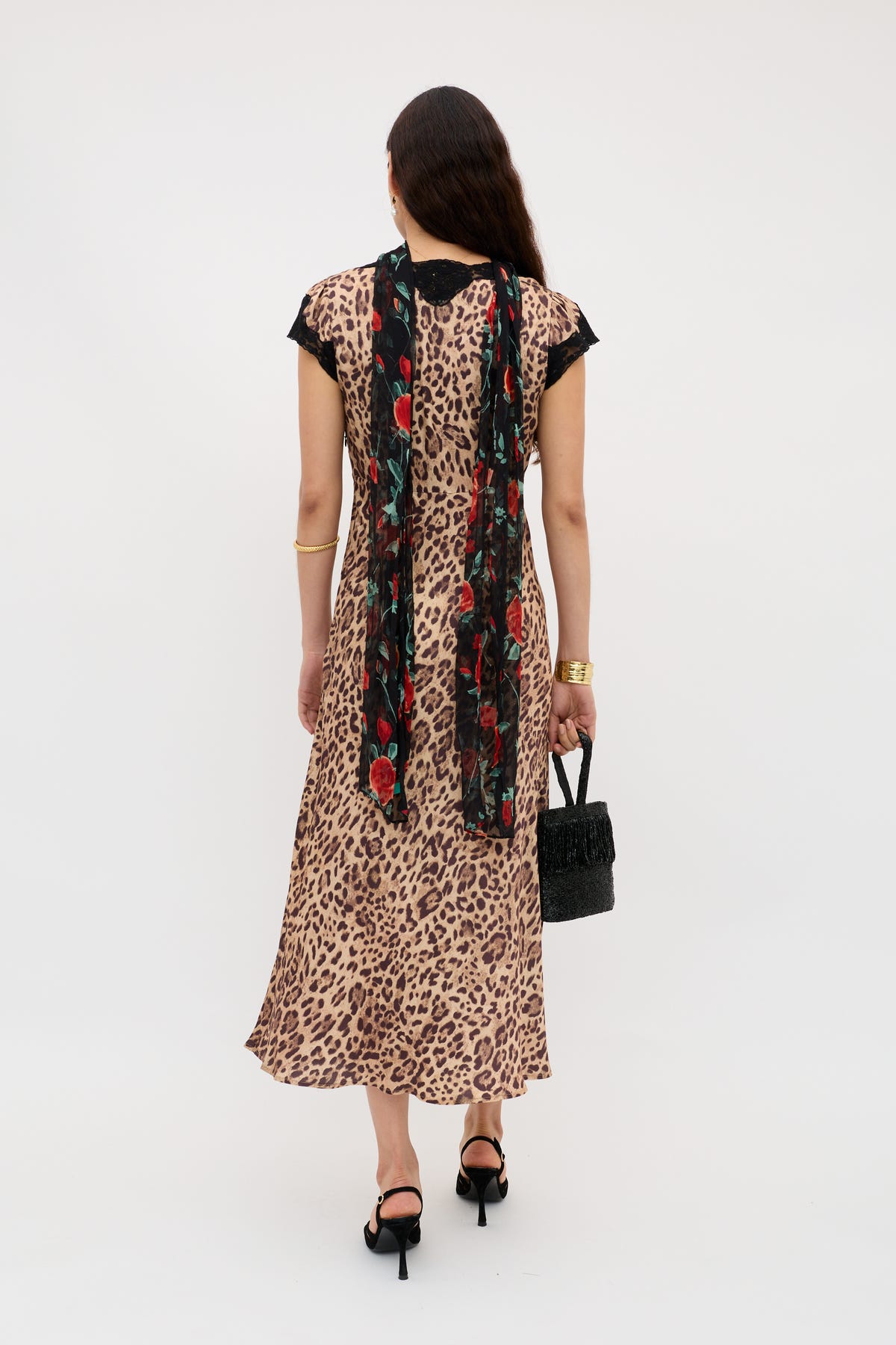 A woman stands facing away in the Amari - Mila Leopard Brown calf-length bias cut dress, black heeled sandals, a black floral scarf with red accents, holding a black textured handbag and wearing gold jewelry.
