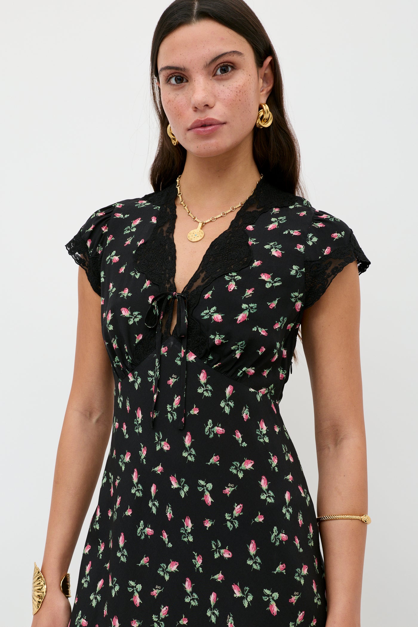 A woman with long brown hair wears the Amari - Sienna Bud Black dress, accessorized with gold jewelry, standing against a plain white background.