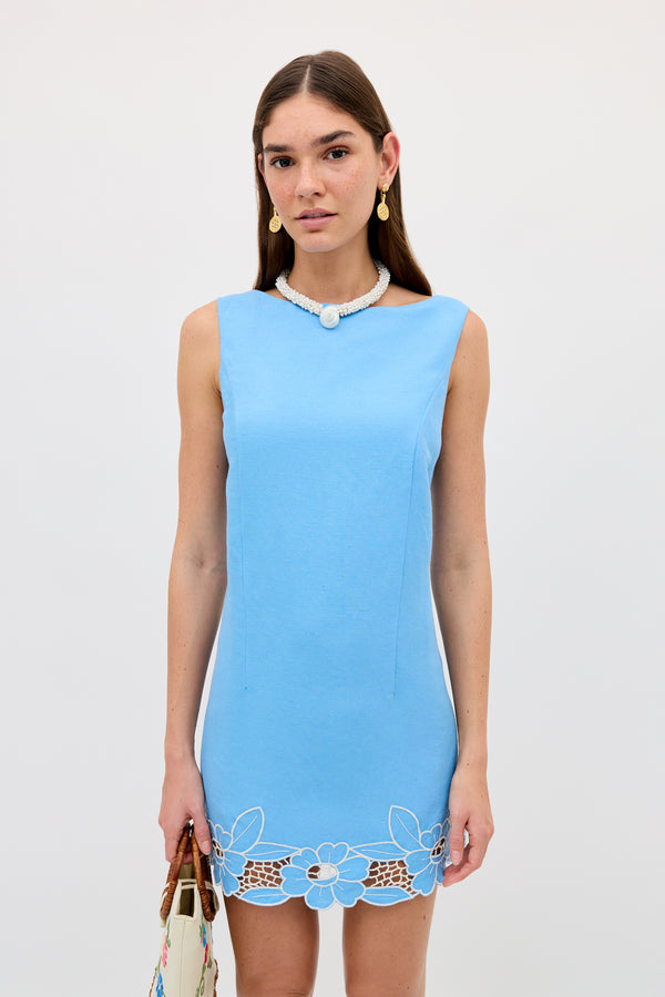 A woman wears the Bingley - Daisy Cutwork Embroidery Blue sleeveless linen dress, paired with a beaded necklace and gold earrings, standing against a white background while holding a white bag.