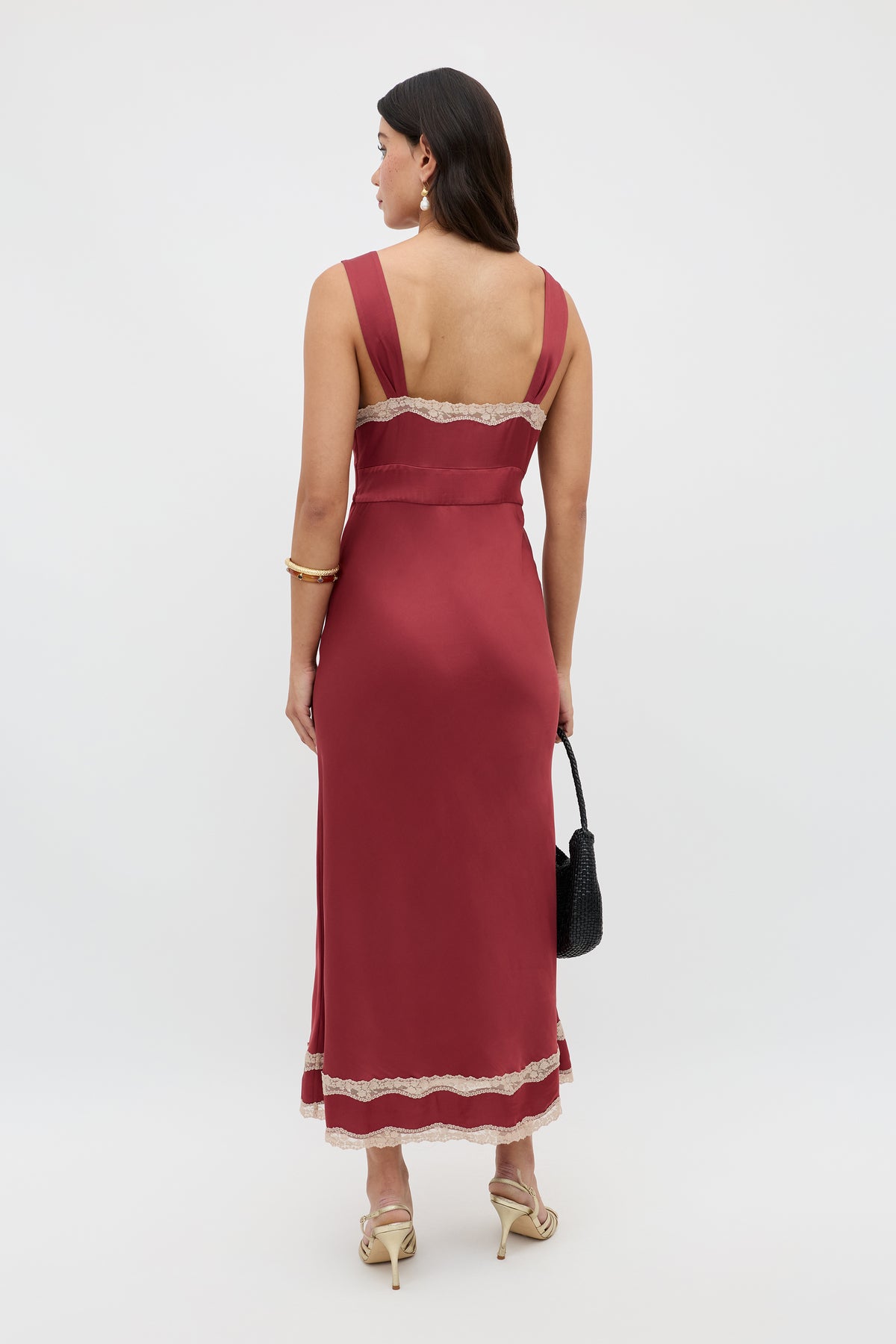 A woman stands facing away, wearing the Lucie - Rose sleeveless, ankle-length burgundy dress with lace trim, beige heels, a black handbag, gold hoop earrings and bracelet, her long dark hair draped down her back.