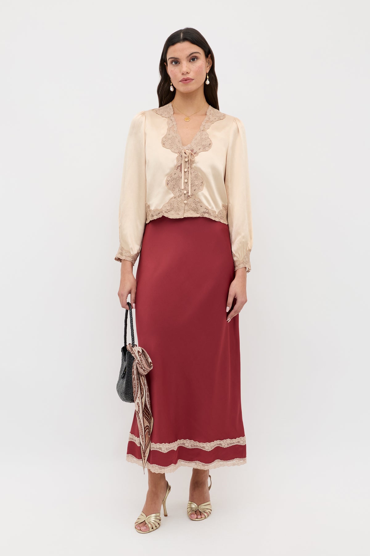 A woman stands against a white background wearing the Lucie - Rose: a cream satin blouse with lace trim and a long burgundy skirt with lace details. She holds a black handbag with a patterned scarf and wears gold heels.