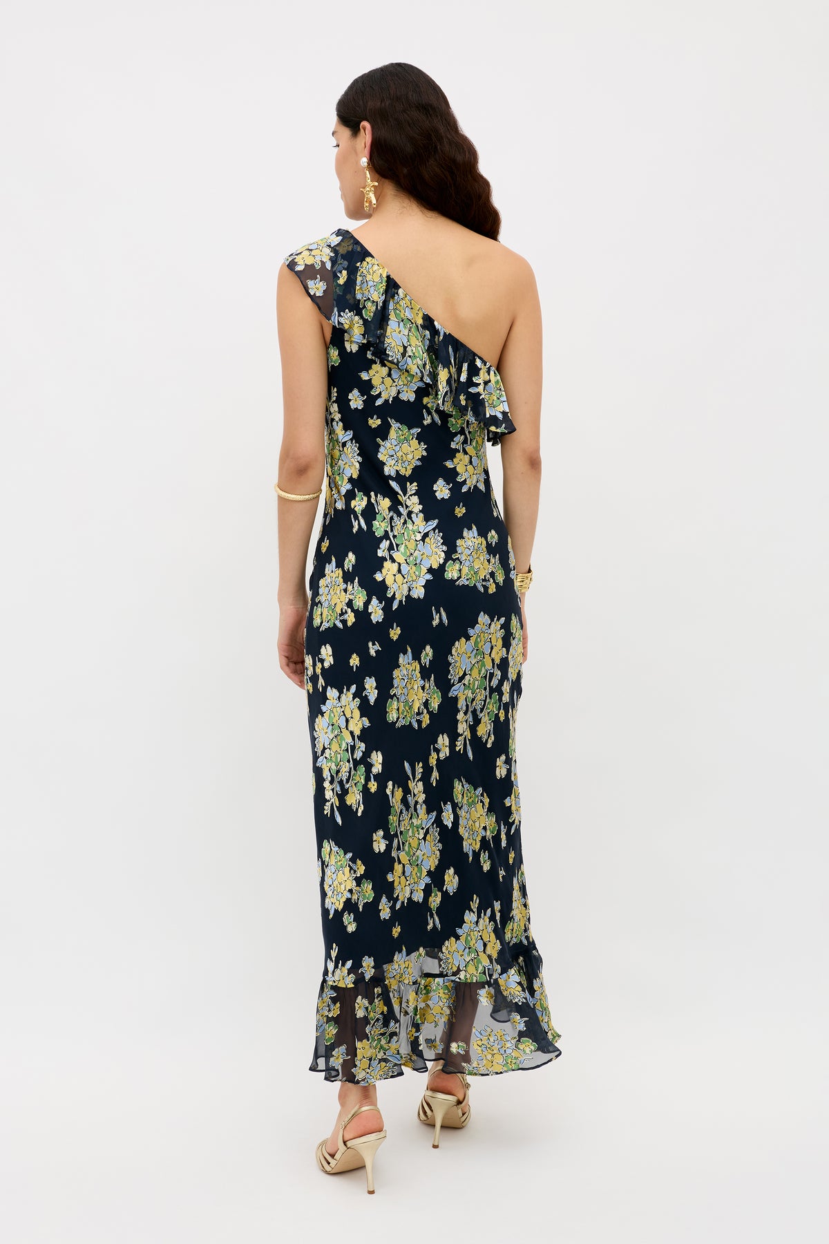 A woman faces away in the Leon - Bouquet Devoré Navy, a black one-shoulder, ankle-length dress with yellow and white floral patterns. She wears gold jewelry and beige heels, standing against a plain white background.
