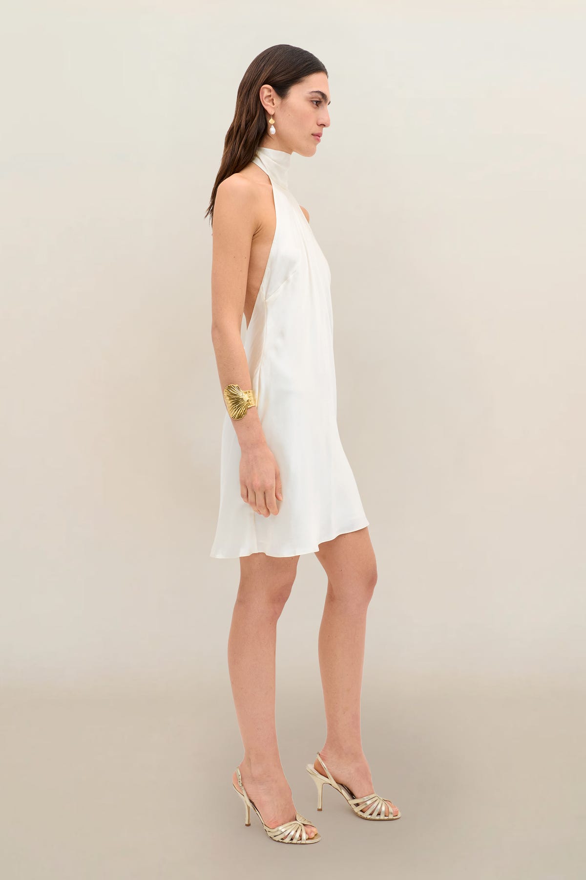 Wearing the Capucine - Champagne, a woman stands sideways against a plain beige background. The sleeveless, halter-neck party dress in lustrous champagne silk-satin features an open back, paired with heeled sandals. She completes her look with a large bracelet and drop earrings.