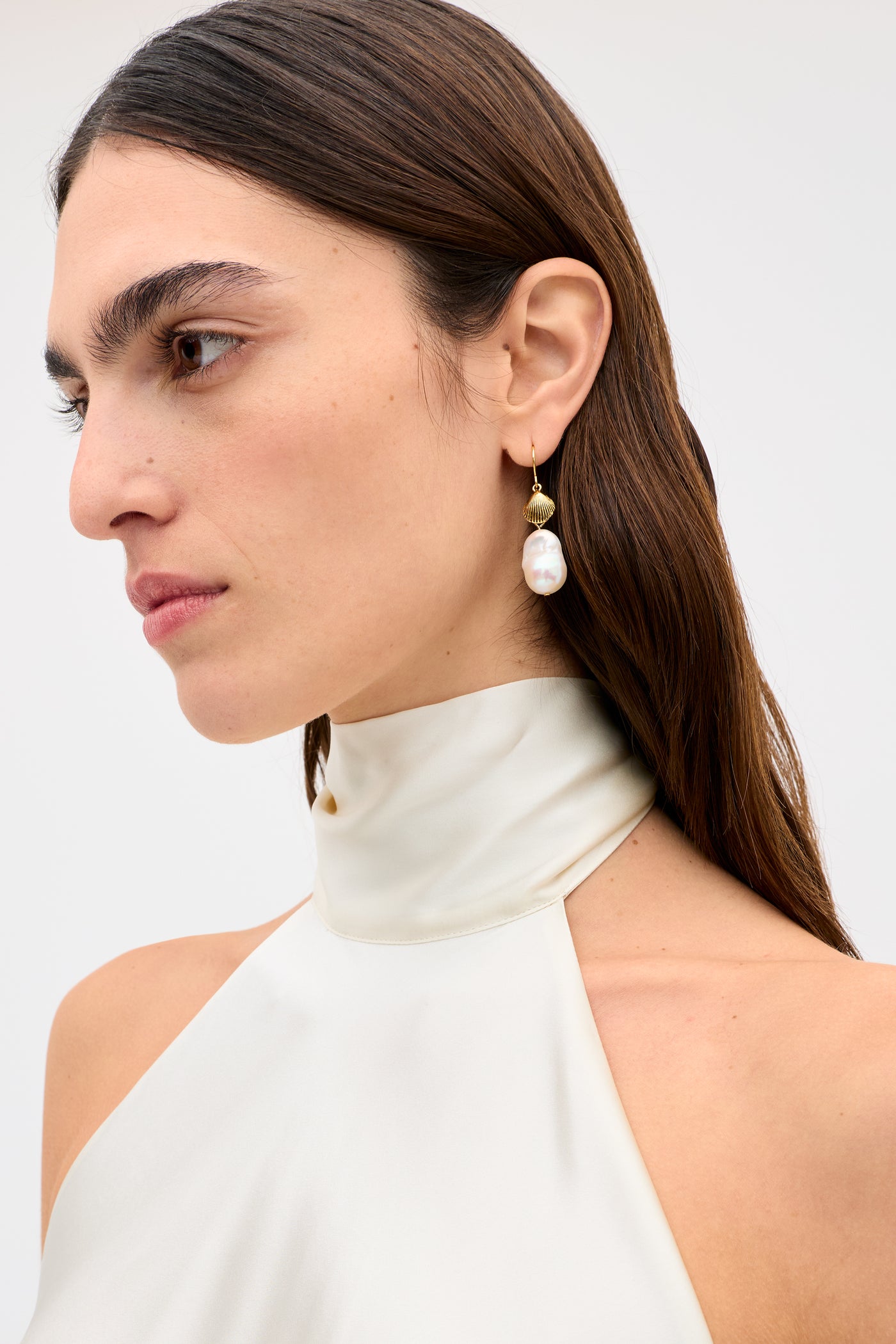 A woman with long brown hair wears the Capucine - Champagne cream high-neck silk-satin top and a gold earring with a large pearl, looking left against a plain light background.
