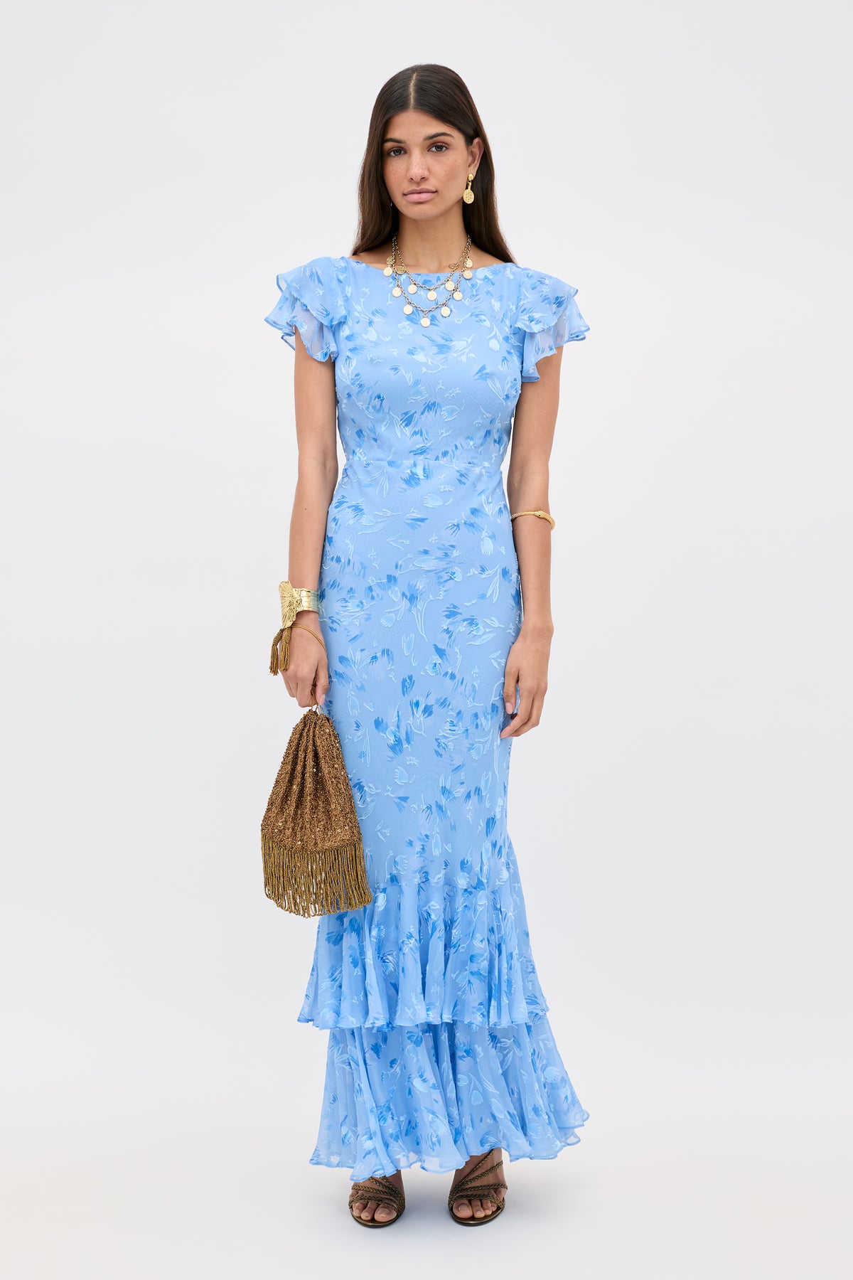 A woman stands facing forward against a plain white background, wearing the Ordinia - Ava Devore Blue dress, accessorized with gold jewelry and holding a brown textured handbag.