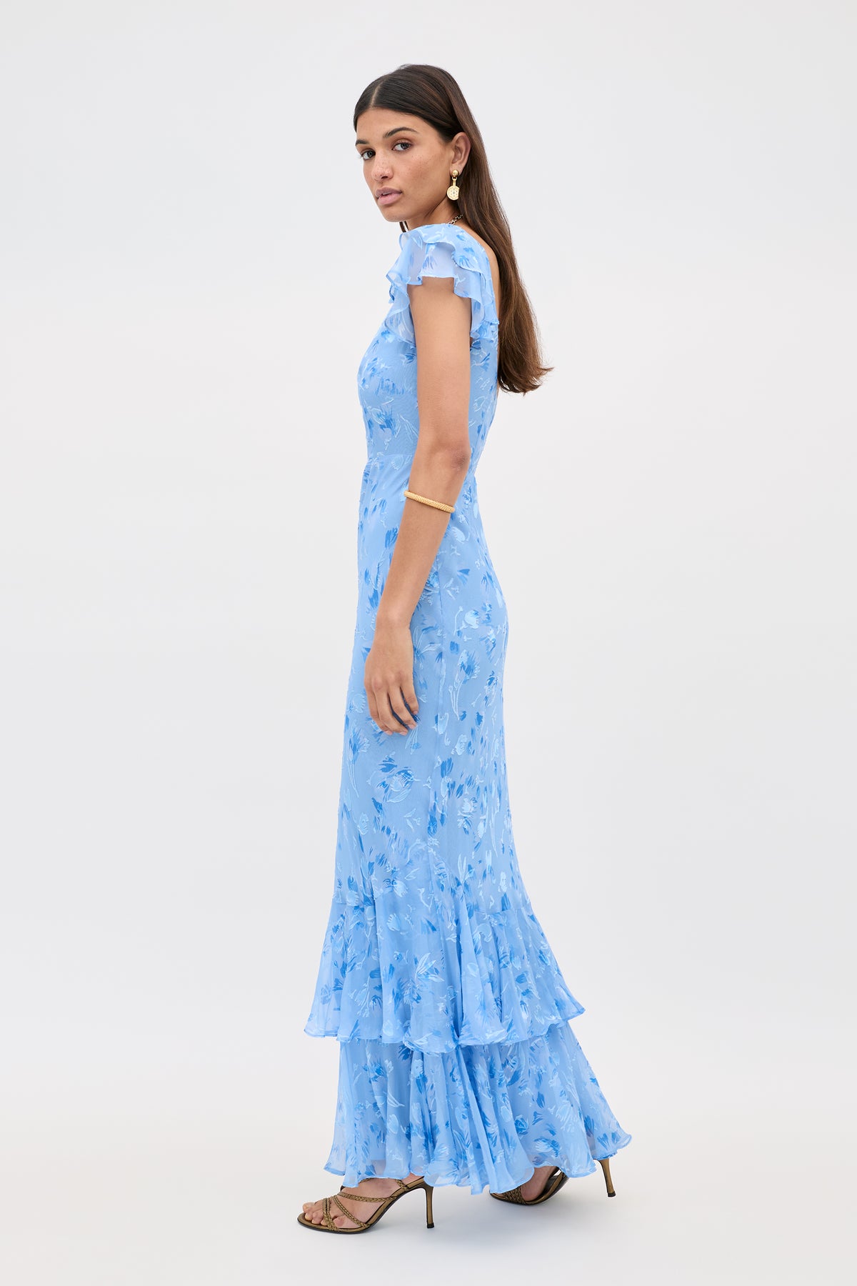 A woman in profile wears the Ordinia - Ava Devore Blue, a light blue floral maxi dress with short sleeves and ruffled hem, paired with gold earrings and strappy heels, standing against a plain white background.