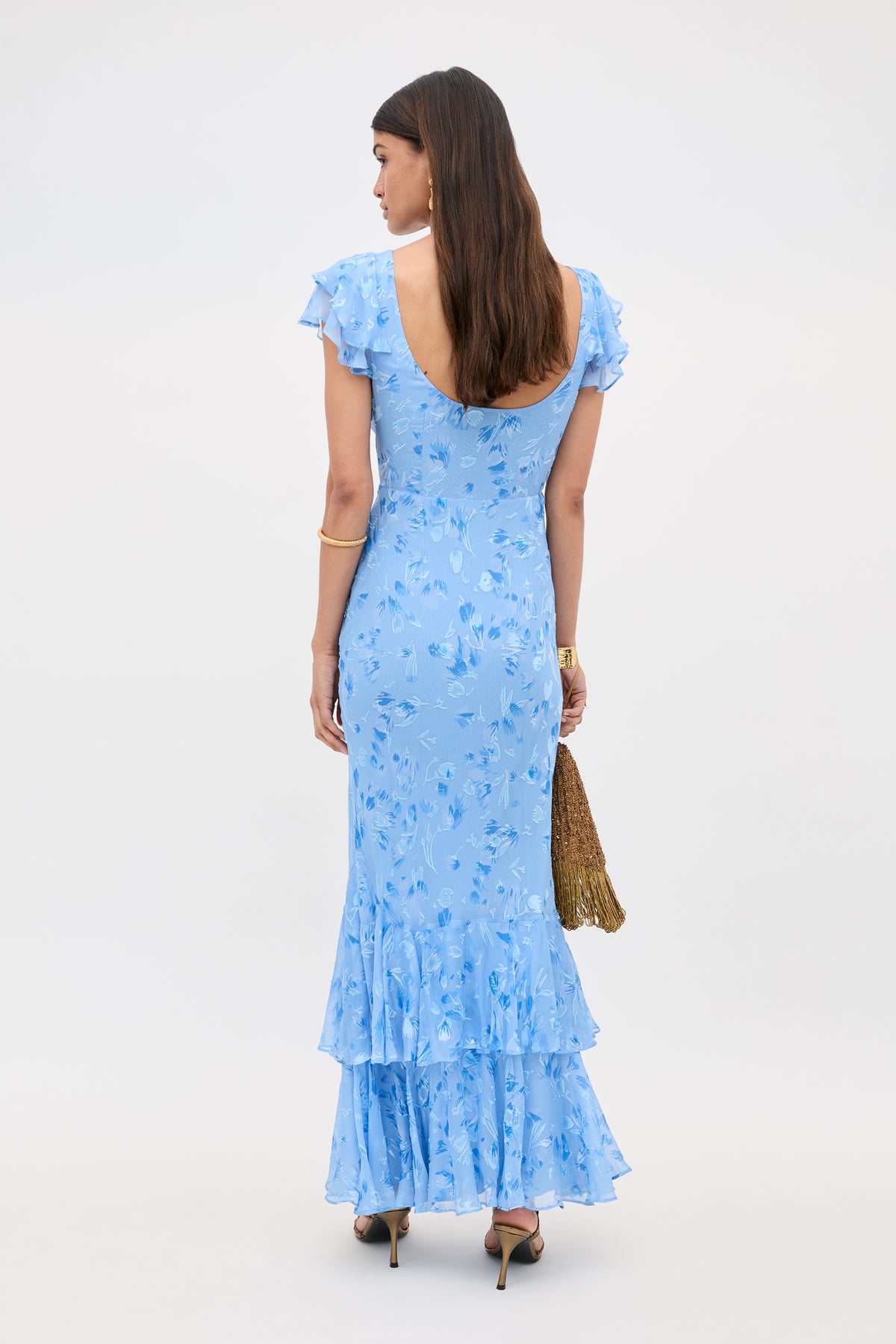 A woman with long brown hair faces away, wearing the Ordinia Ava Devore Blue—a light blue, floral ruffled dress with short sleeves and a scoop back. She holds a gold tassel bag and wears high heels against a plain, light background.