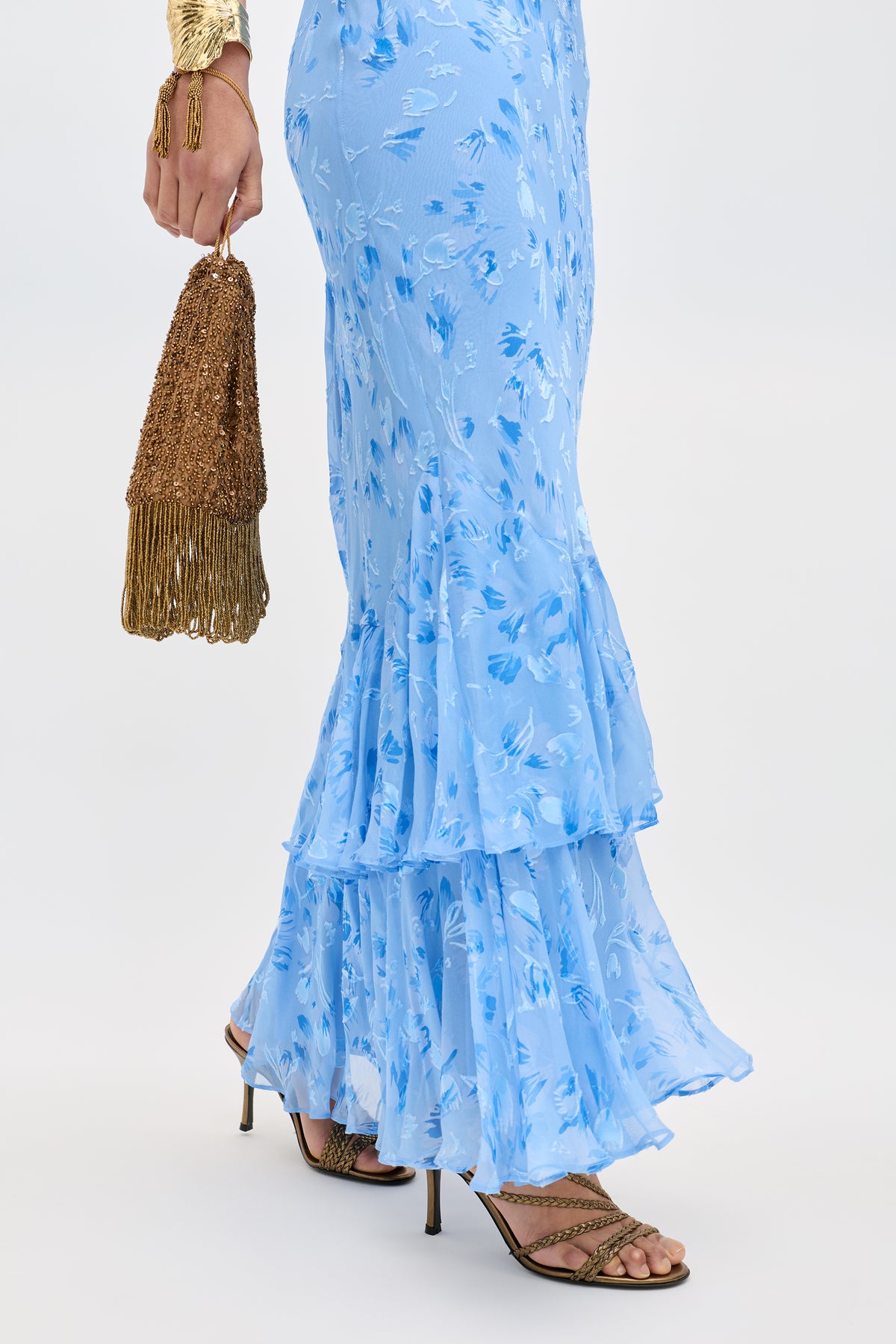 A person wearing the Ordinia Ava Devore Blue floral ruffled dress with strappy heeled sandals holds a gold beaded fringe handbag. Only their lower body and hand are visible against a plain background.