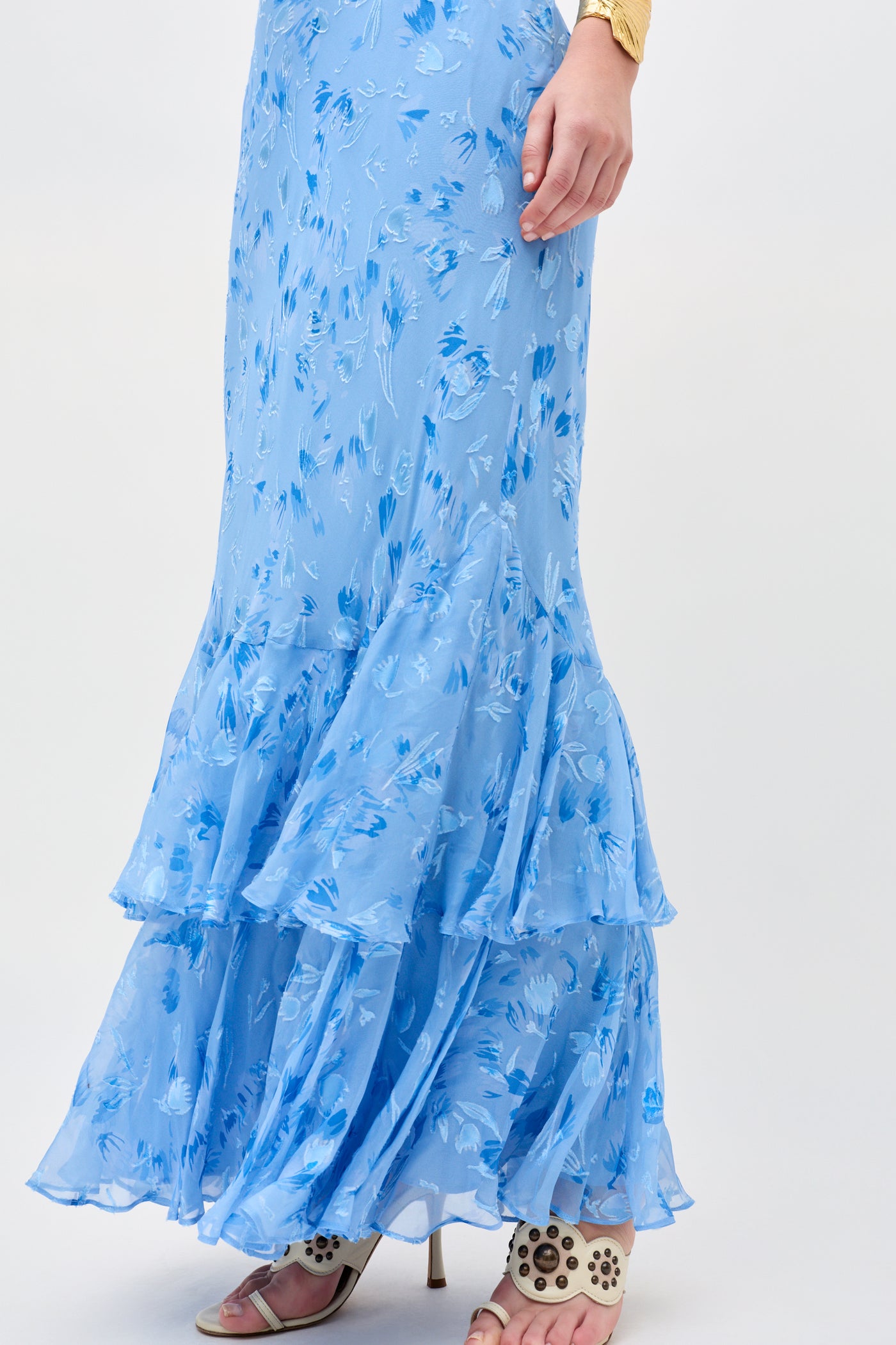 Wearing the Ordinia - Ava Devore Blue, a flowing light blue floral ruffled skirt, paired with cream cut-out high heels. Their hand rests casually at their side.
