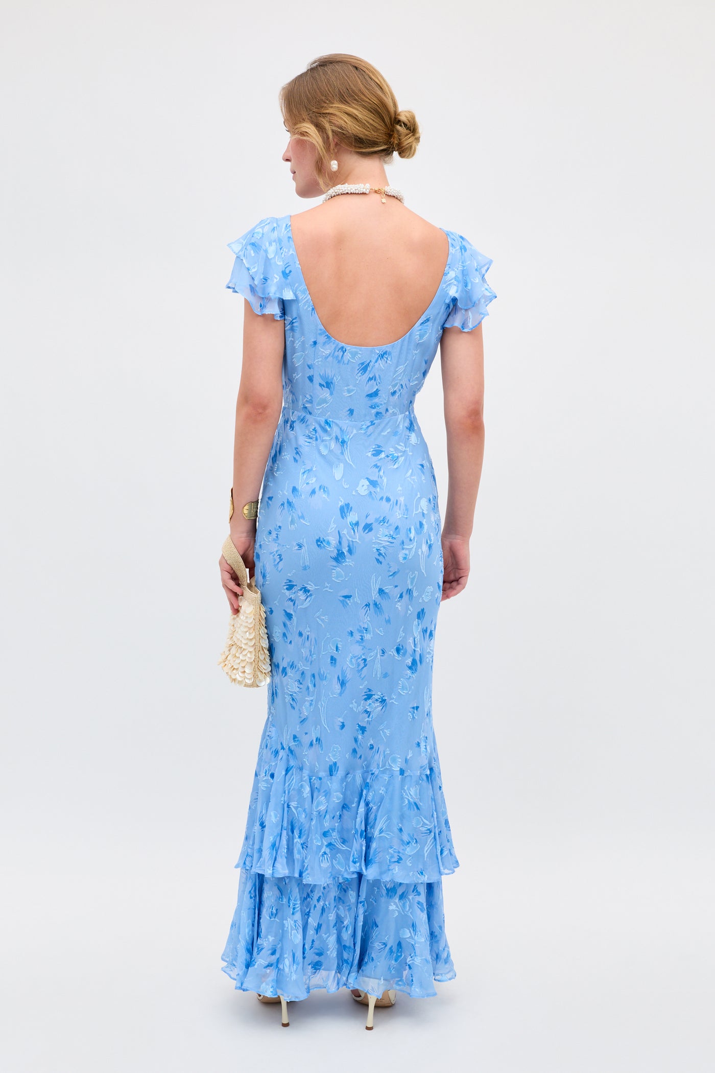 A woman faces away in the Ordinia Ava Devore Blue dress, a light blue, floral-patterned, floor-length gown with ruffled sleeves and low back. She carries a beaded handbag, wears heels, pearl necklace, bracelet, and her hair is in a low bun.