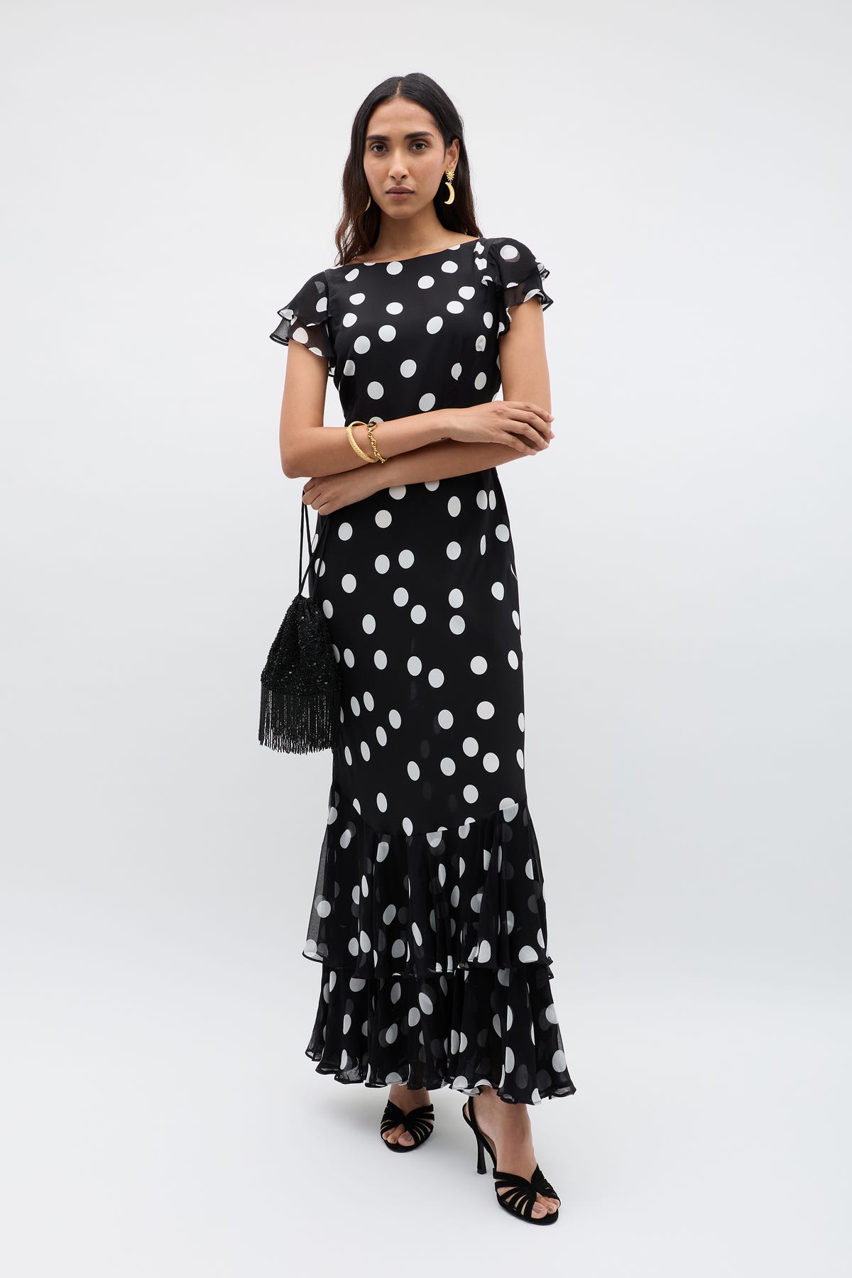 A woman wears the Ordinia – Mono Polka Dot Black, a vintage-inspired ankle-length formal dress with white polka dots, ruffled hem, and flutter sleeves. She pairs it with a black fringe bag, black heels, gold earrings, and a bracelet.