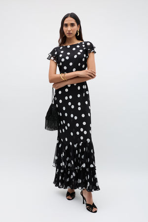 A woman wears the Ordinia – Mono Polka Dot Black, a vintage-inspired ankle-length formal dress with white polka dots, ruffled hem, and flutter sleeves. She pairs it with a black fringe bag, black heels, gold earrings, and a bracelet.